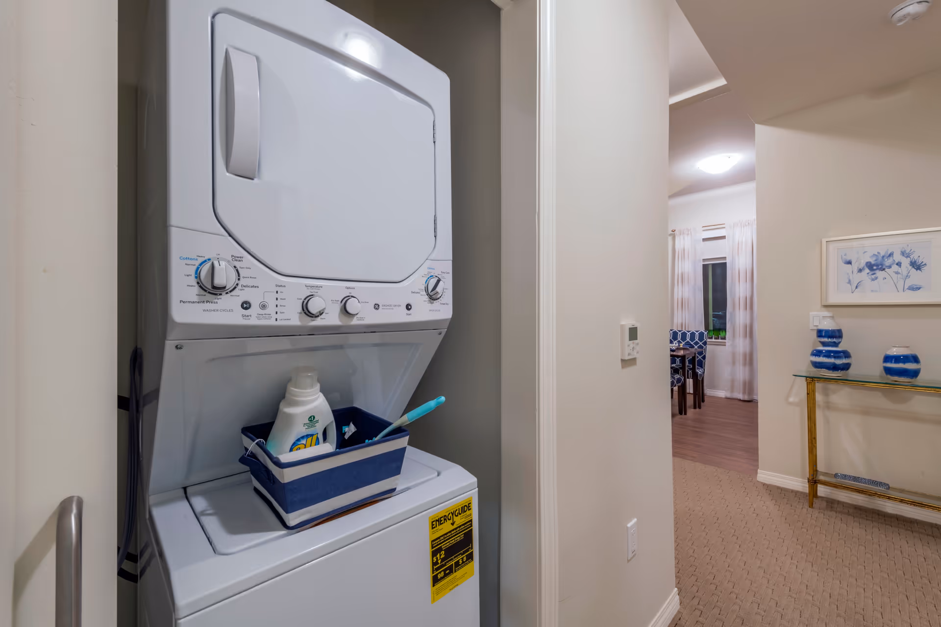 Stacked washer and dryer in a hallway laundry closet with a detergent basket and a view into a furnished living/dining area.