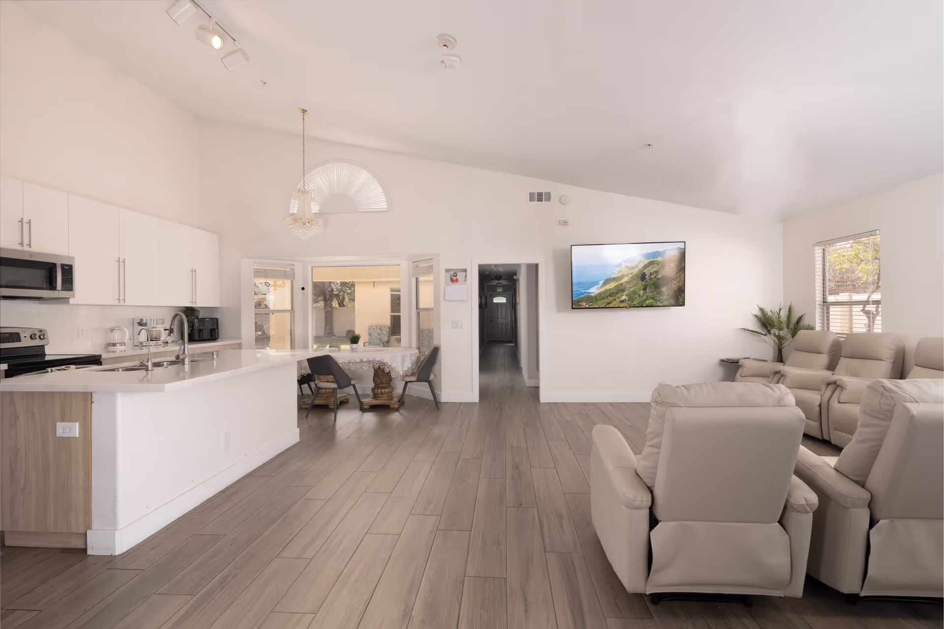 Bright and spacious interior of an assisted living facility featuring a modern kitchen with white cabinets and stainless steel appliances on the left, a small dining area with a table and chairs near a window in the center, and a living room area with beige recliner chairs facing a wall-mounted flat-screen TV on the right. The room has light wood flooring and white walls with large windows allowing natural light.