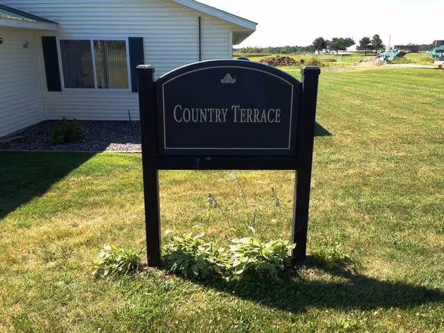 A black sign with white text reading 'Country Terrace' stands on a grassy lawn in front of a light-colored building with a window and dark shutters. There are small plants at the base of the sign and open grassy fields in the background under a clear sky.