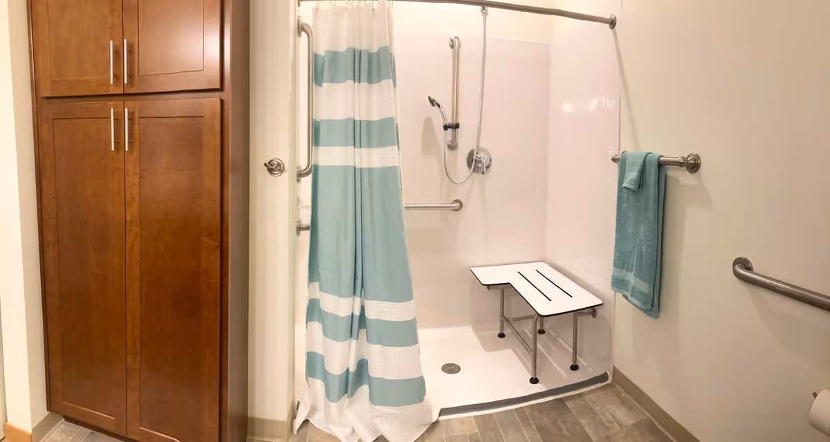 A senior-friendly bathroom featuring a walk-in shower with a white and teal striped curtain, a foldable shower bench, grab bars on the walls, a handheld showerhead, a teal towel hanging on a towel bar, and a wooden cabinet for storage.
