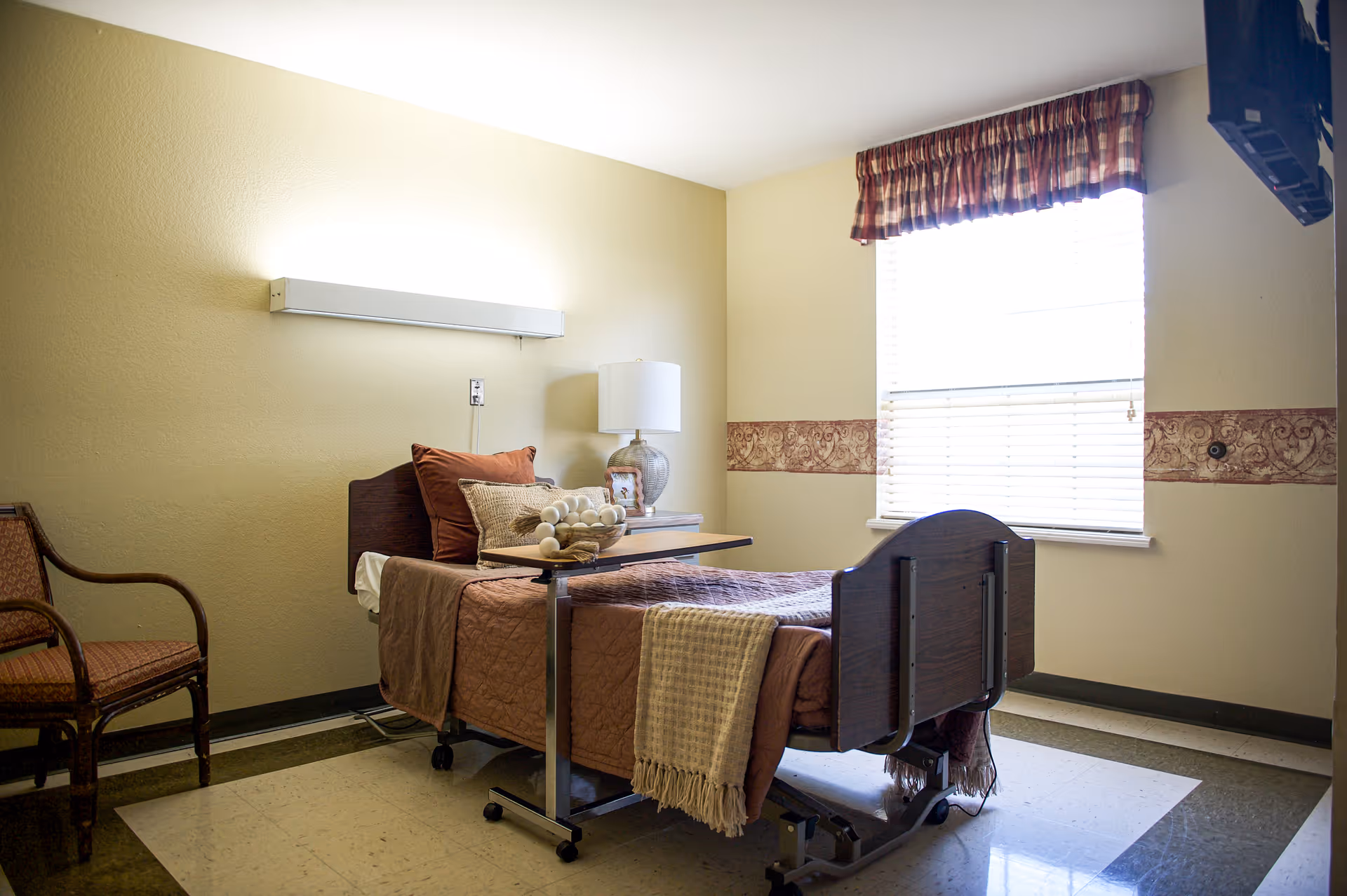A well-lit senior facility bedroom with a hospital-style bed, bedside table, lamp and chair by a window.