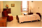 A simple furnished bedroom with a single bed covered by a floral quilt, a recliner, dresser, side table with lamp, and a window with patterned curtains.