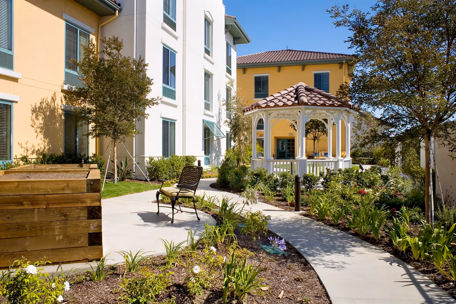 Outdoor garden area at Belmont Village Thousand Oaks featuring a curved concrete pathway, a wooden planter box, a metal chair with a cushion, various plants and flowers, trees, and a white gazebo with a tiled roof adjacent to a yellow and white building under a clear blue sky.