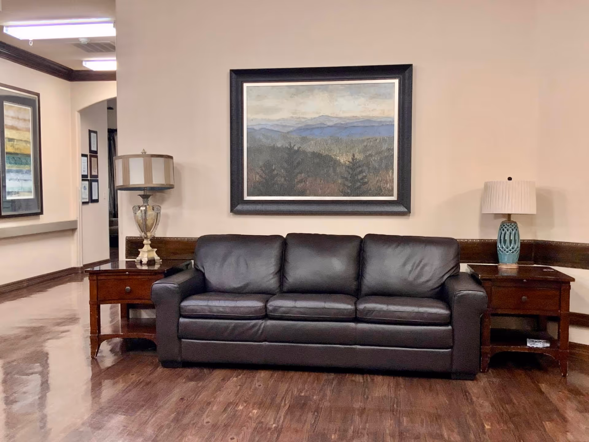 A cozy interior seating area featuring a dark brown leather sofa centered between two wooden side tables, each with a decorative lamp. Above the sofa hangs a framed landscape painting depicting mountains and trees. The room has polished wooden floors and beige walls with dark wood trim.