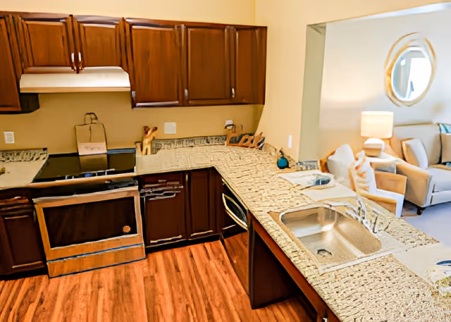 A kitchen area with wooden cabinets, a stove, a dishwasher, and a granite countertop with a sink. The kitchen opens into a living room area with a beige armchair, a side table with a lamp, and a round mirror on the wall.