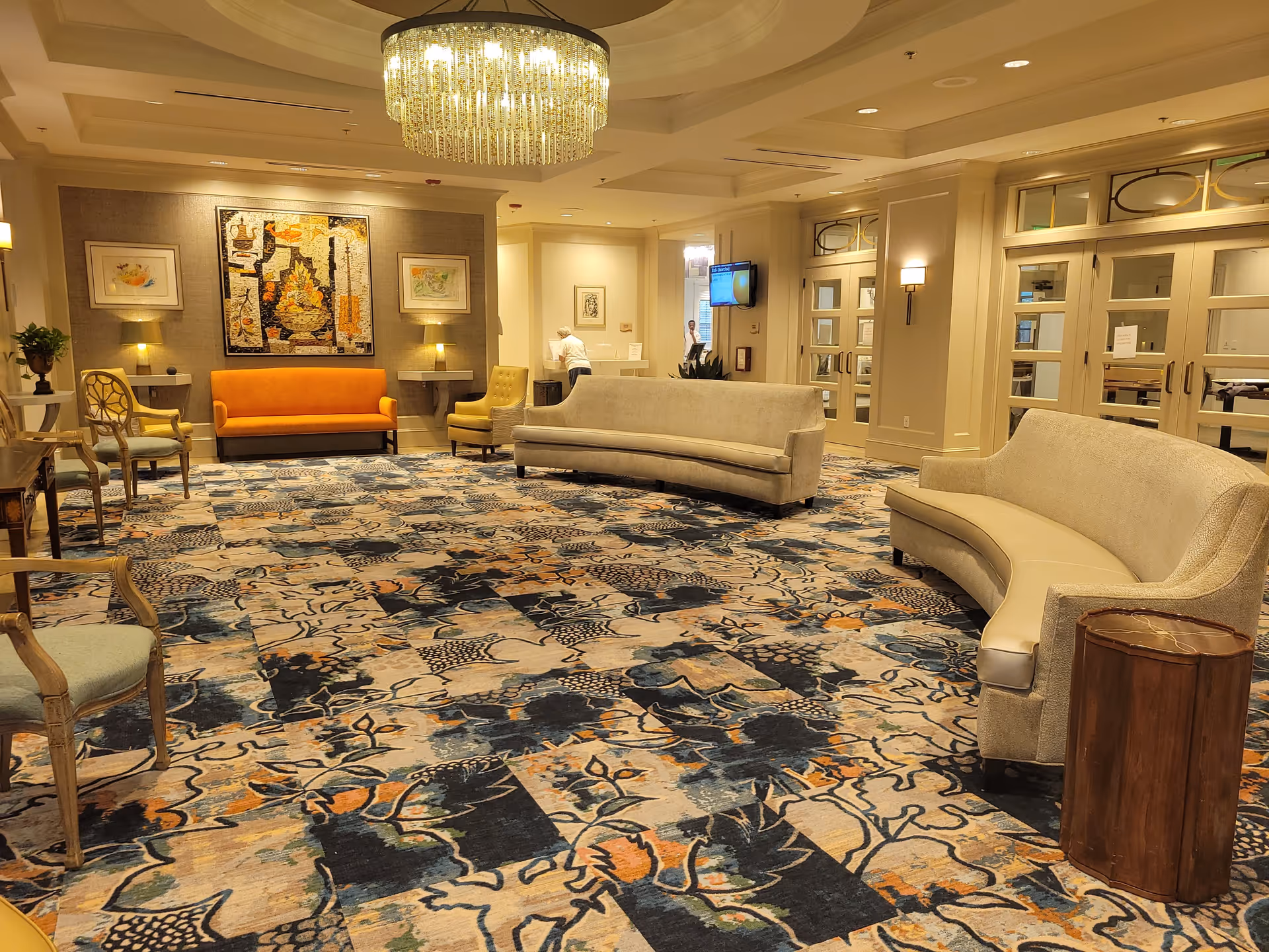 A spacious, well-lit lounge area with a patterned carpet in shades of blue, beige, and orange. The room features two curved beige sofas, an orange sofa against the far wall, several chairs, and small tables. A large chandelier hangs from the ceiling, and framed artwork is displayed on the walls. There are double glass doors on the right side and a person is visible in the background near a reception or service desk.