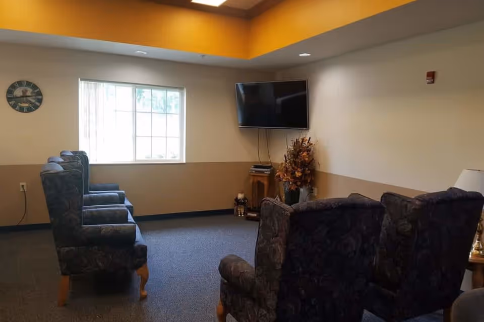 A cozy senior living common area with several upholstered armchairs arranged facing a wall-mounted flat screen TV. There is a window letting in natural light, a clock on the wall, and a decorative plant near the TV. The walls are painted beige with a darker beige accent near the ceiling.
