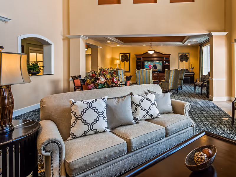 A cozy living room area with a beige sofa adorned with patterned and plain cushions. In front of the sofa is a dark wooden coffee table with decorative items. The room has warm lighting, a patterned carpet, and a lamp on a side table. In the background, there is a dining area with chairs and a wooden entertainment center with a TV and decorative wall art.