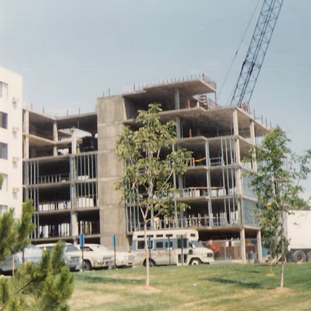 Partially constructed multi-story concrete building with a crane, parked vehicles, and trees on a grassy foreground.