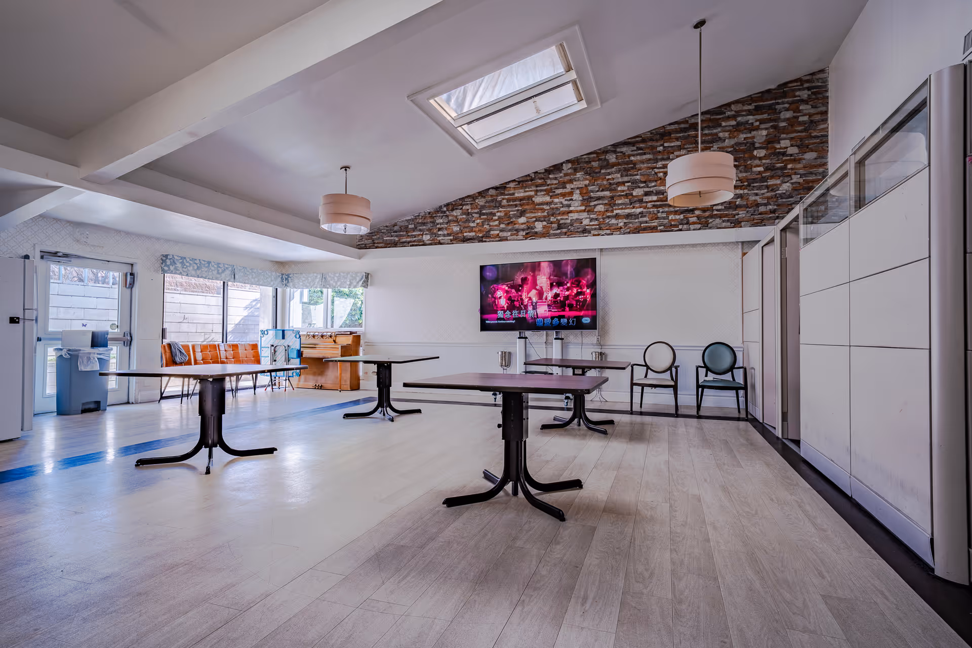 A spacious room with light wood flooring and a high ceiling featuring a skylight. The room has three tables with black bases and dark tops spaced apart. On one side, there is a wall-mounted flat-screen TV displaying a colorful scene with text. Two chairs with round backs are placed against the wall beneath the TV. The opposite side has large windows with a door, allowing natural light to enter. There is a bench with orange cushions and a foosball table near the windows. The ceiling has two pendant lights with beige lampshades, and one wall is decorated with a stone brick pattern.