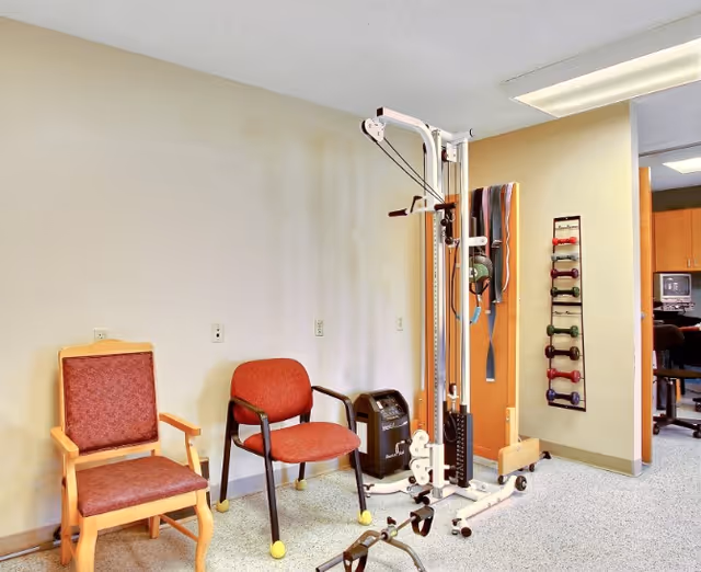 Small therapy/exercise room with two chairs, a cable exercise machine, and a wall-mounted rack of hand weights.