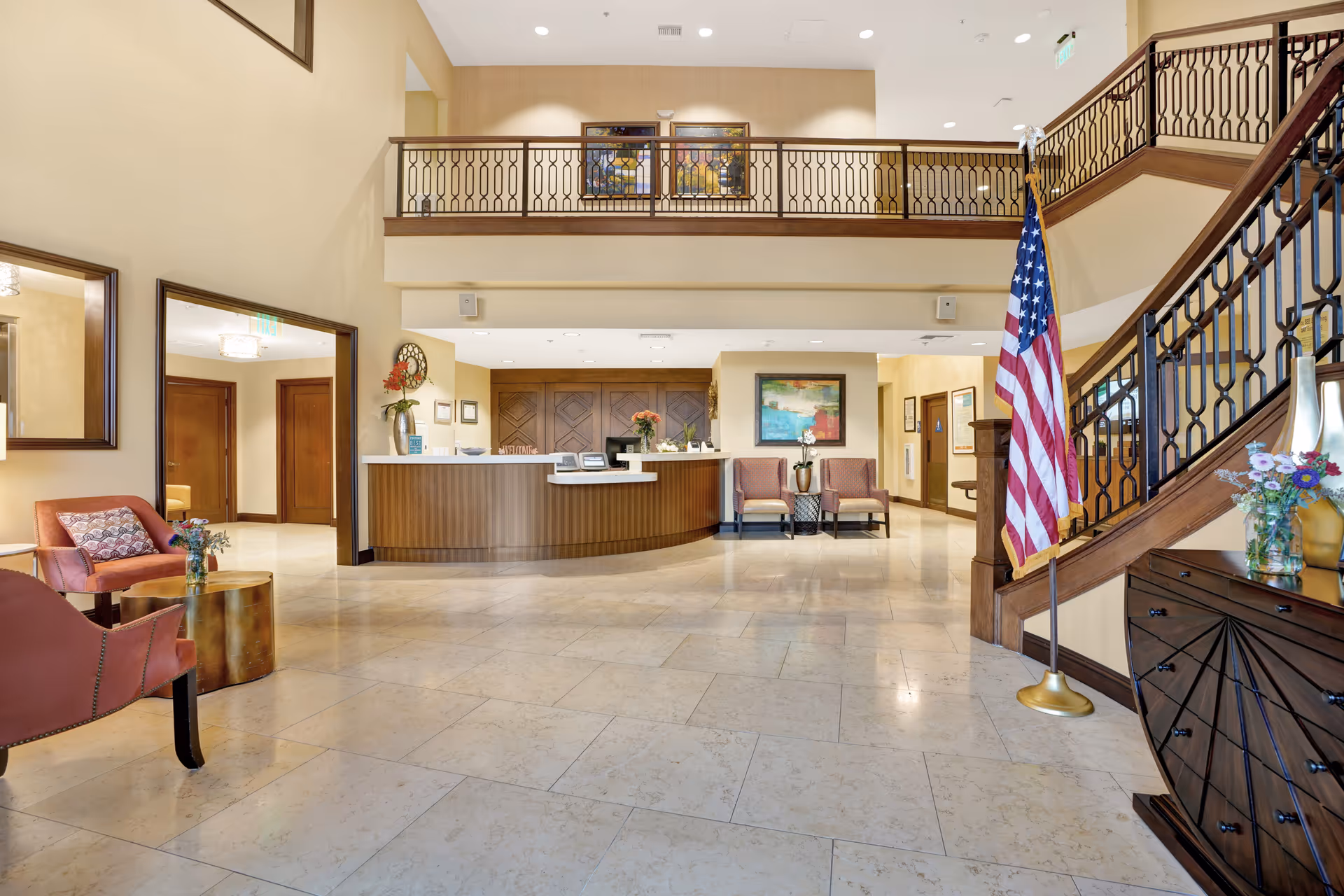 Spacious and well-lit senior living facility lobby with a reception desk in the center, two chairs and a small table with flowers on the left, an American flag near a staircase with decorative railing on the right, and artwork on the walls.