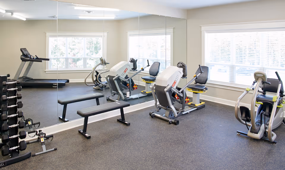 A well-lit fitness room with exercise equipment including a treadmill, weight bench, dumbbells, and various strength training machines. Large windows allow natural light to fill the room, and a wall-sized mirror reflects the equipment.