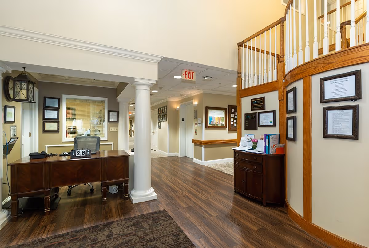 Interior view of a senior living facility reception area with a wooden desk, office chair, and framed certificates on the walls. The space features wooden flooring, white columns, a staircase with wooden railing, and an exit sign on the ceiling.