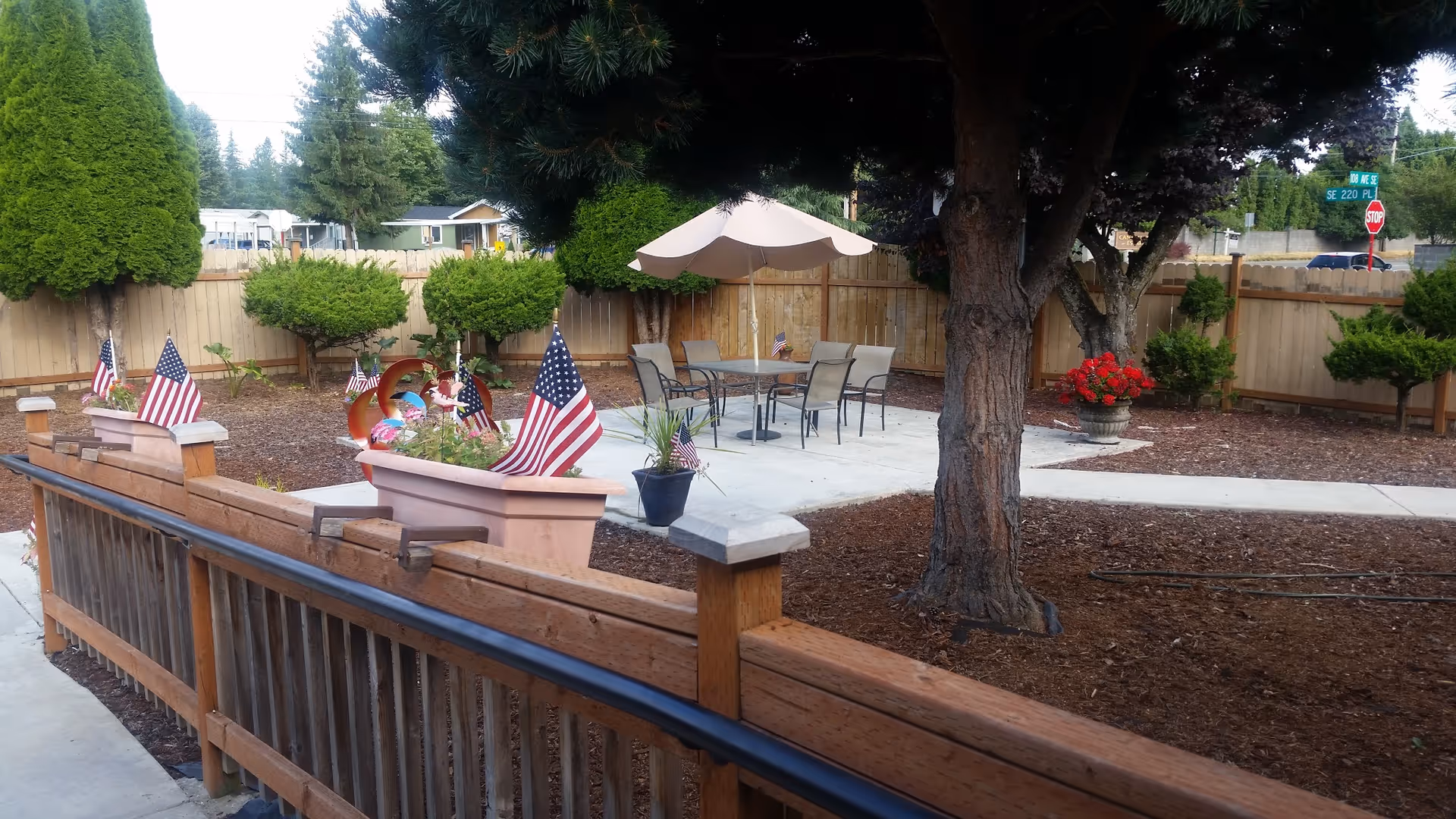 Outdoor courtyard with a patio table and umbrella, potted plants and American flags, surrounded by trees and a wooden fence.