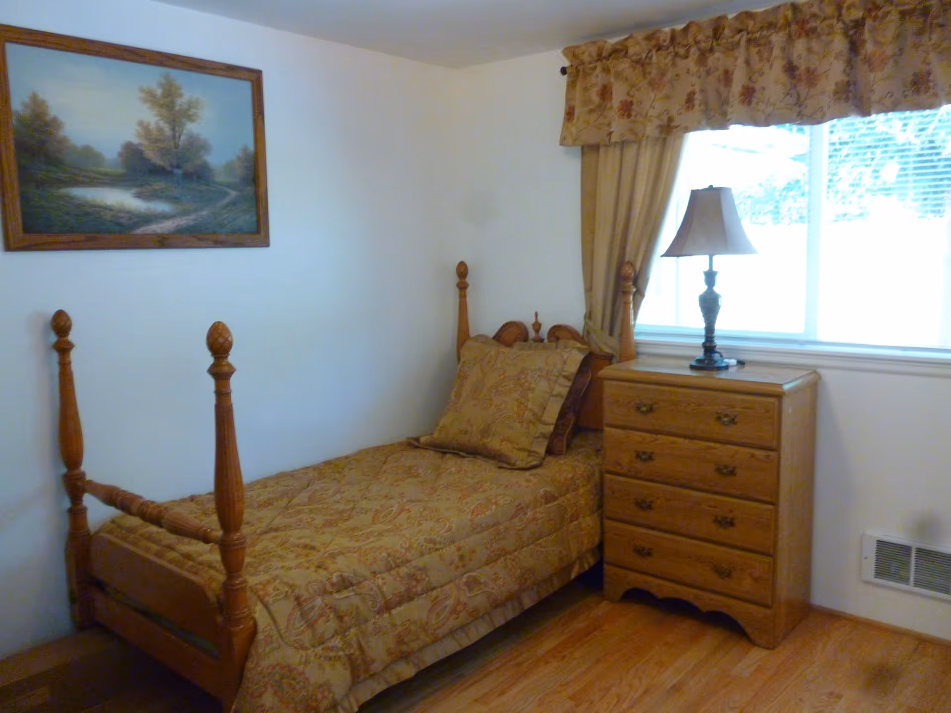 Small single bedroom with a wooden four-poster bed, patterned bedding, a dresser with lamp, and a window with a valance.