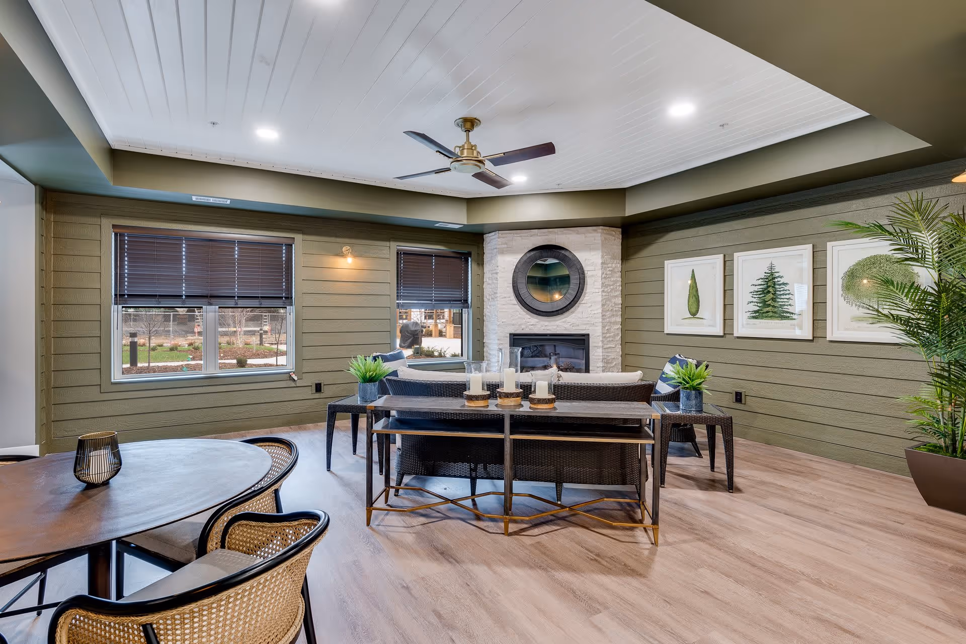 A cozy living room area with green paneled walls, a white ceiling with recessed lighting, and a ceiling fan. The room features a stone fireplace with a round mirror above it, a wicker sofa and chairs with cushions, a wooden table with candles, and framed botanical artwork on the wall. There are two windows with dark blinds and a potted plant in the corner.