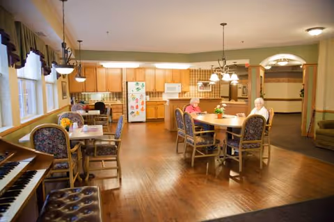 A spacious dining area in a senior living facility with wooden floors and multiple tables and chairs. Two elderly women are seated at a round table near the kitchen area, which features wooden cabinets, a refrigerator with colorful magnets, a microwave, and a stove. The room is well-lit with hanging light fixtures and large windows with curtains. A piano is visible in the foreground.