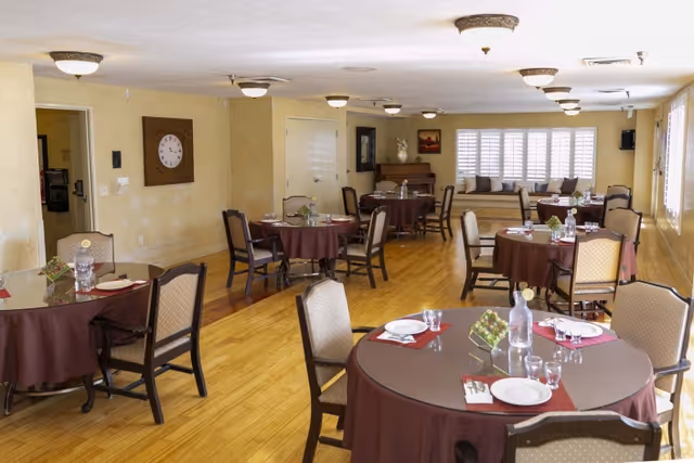 A dining room with several round tables covered with brown tablecloths, each set with plates, glasses, and utensils. The room has wooden flooring, beige walls, ceiling lights, and a window with white shutters. There are cushioned chairs around the tables and a piano against the far wall.