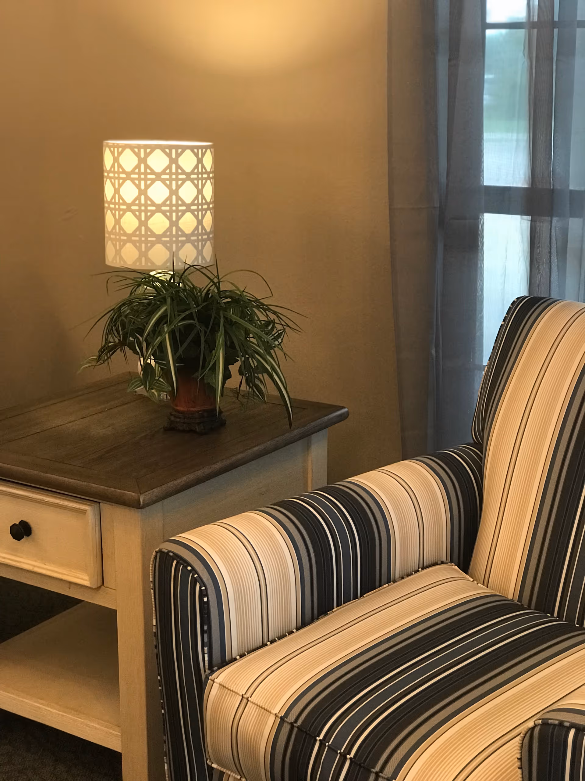 A cozy corner featuring a striped armchair next to a wooden side table with a drawer. On the table, there is a potted green plant and a lit table lamp with a patterned lampshade. A window with sheer curtains is visible in the background.