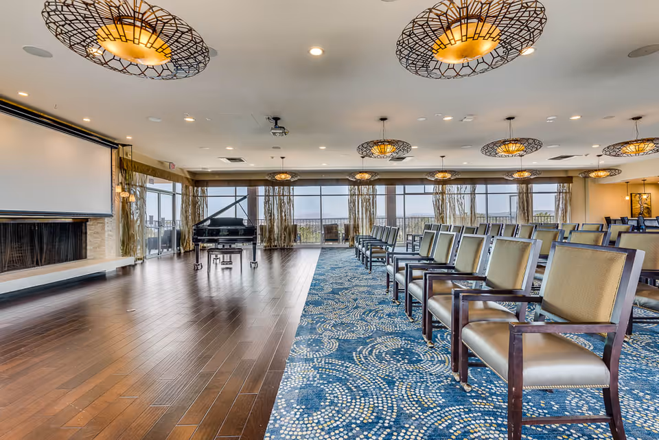 A spacious senior living facility room with rows of chairs arranged facing a large projection screen and a black grand piano. The room features wooden flooring on one side and a blue patterned carpet on the other, with large windows and glass doors letting in natural light. Modern circular light fixtures hang from the ceiling.