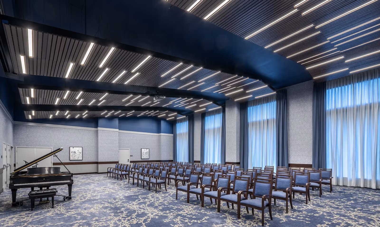 Spacious modern event room with rows of chairs, a grand piano on the left, tall curtained windows, and patterned carpeting.