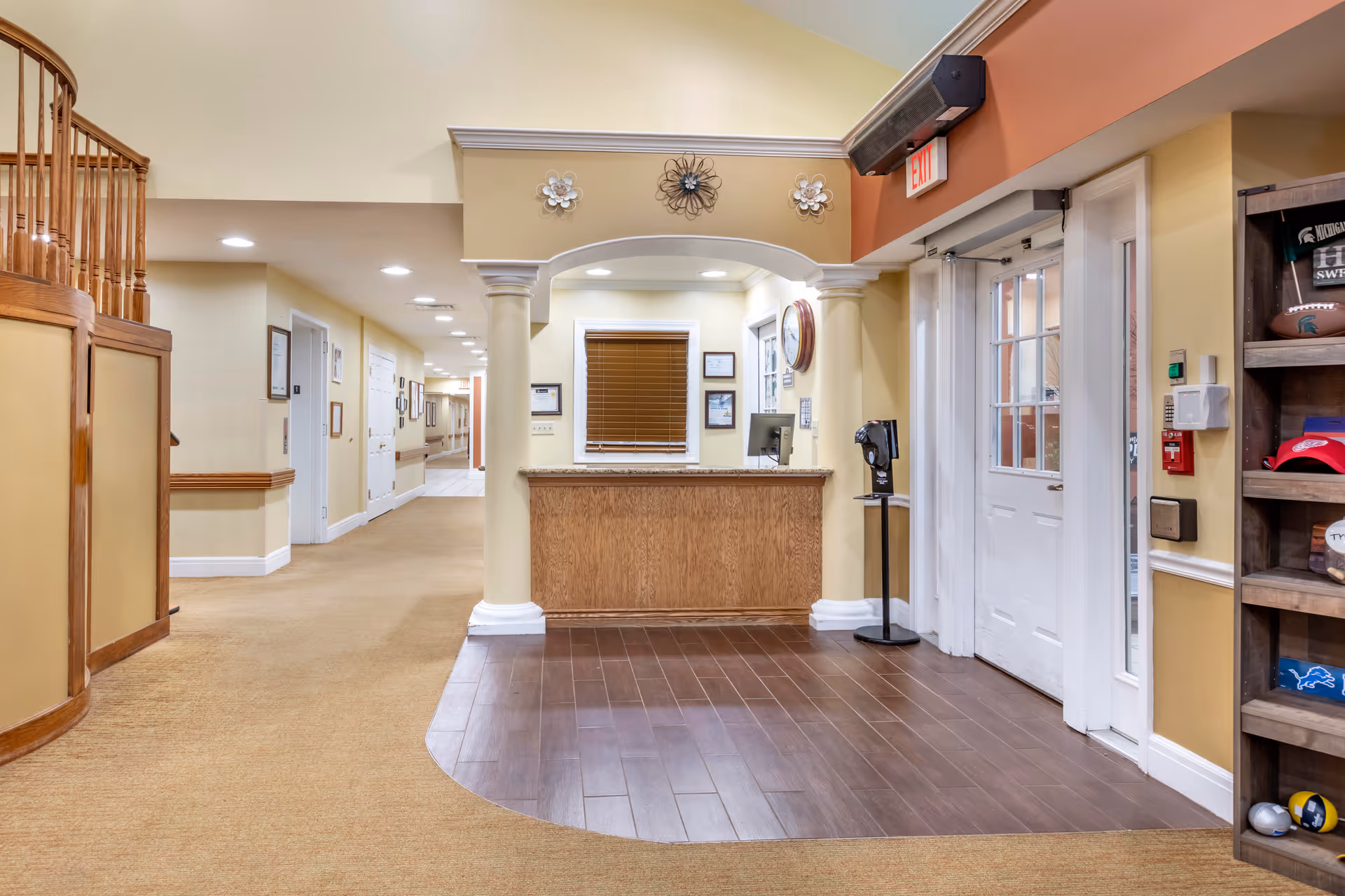 Reception area inside an assisted living facility with a wooden front desk, a computer monitor, and a window with blinds behind it. The floor near the desk is wooden, transitioning to carpet in the hallway. There are decorative wall accents above the desk, an exit door to the right, and a wooden shelf displaying sports memorabilia. The hallway extends to the left with doors and framed pictures on the walls.
