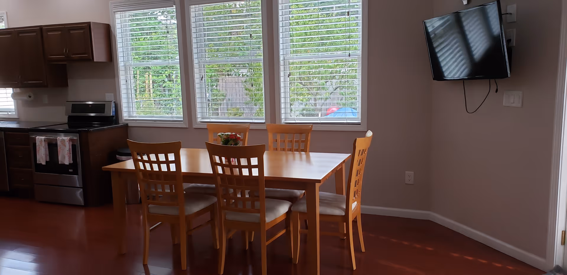 A dining area with a wooden table and six matching chairs placed in front of three large windows with white blinds. To the left, there is a kitchen area with dark wooden cabinets, a stainless steel stove, and a countertop. On the right wall, a flat-screen TV is mounted. The floor is a polished wood finish.