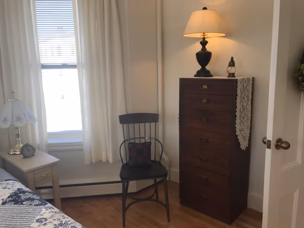 A cozy bedroom corner with a wooden chair featuring a decorative cushion, a tall dark wooden dresser with a lace runner, a table lamp, and a small lantern on top. Next to the dresser is a window with white curtains and blinds, and a bedside table with a floral lamp and an alarm clock is partially visible.