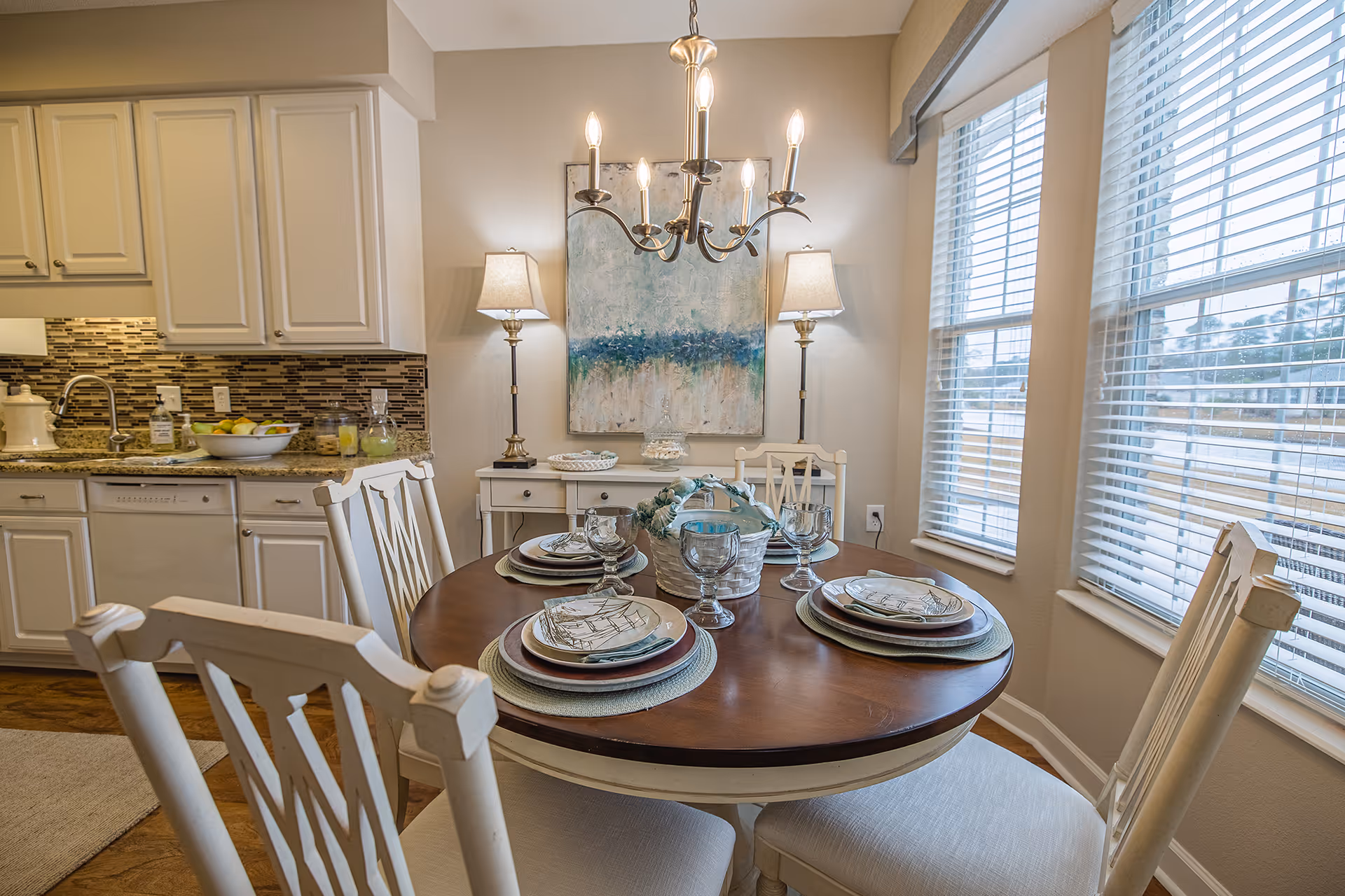 A bright dining area with a round wooden table set for four with plates, glasses, and napkins. The room features white chairs, large windows with blinds letting in natural light, a chandelier overhead, and a sideboard with two lamps and a decorative painting on the wall. The kitchen with white cabinets and a tiled backsplash is visible in the background.