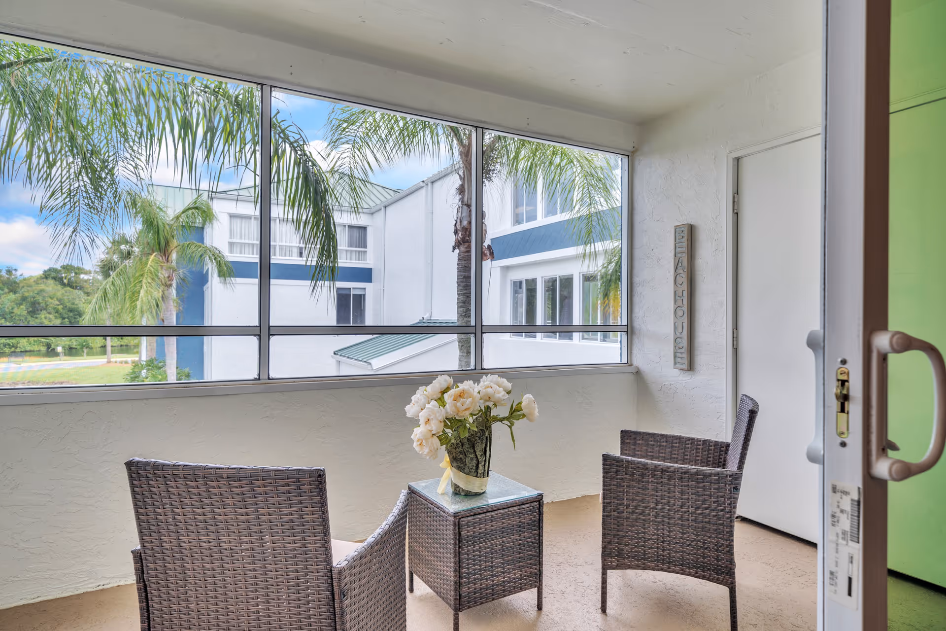 A screened-in balcony with two wicker chairs and a small wicker table holding a vase of white flowers. The balcony overlooks palm trees and a white building with blue accents under a partly cloudy sky.