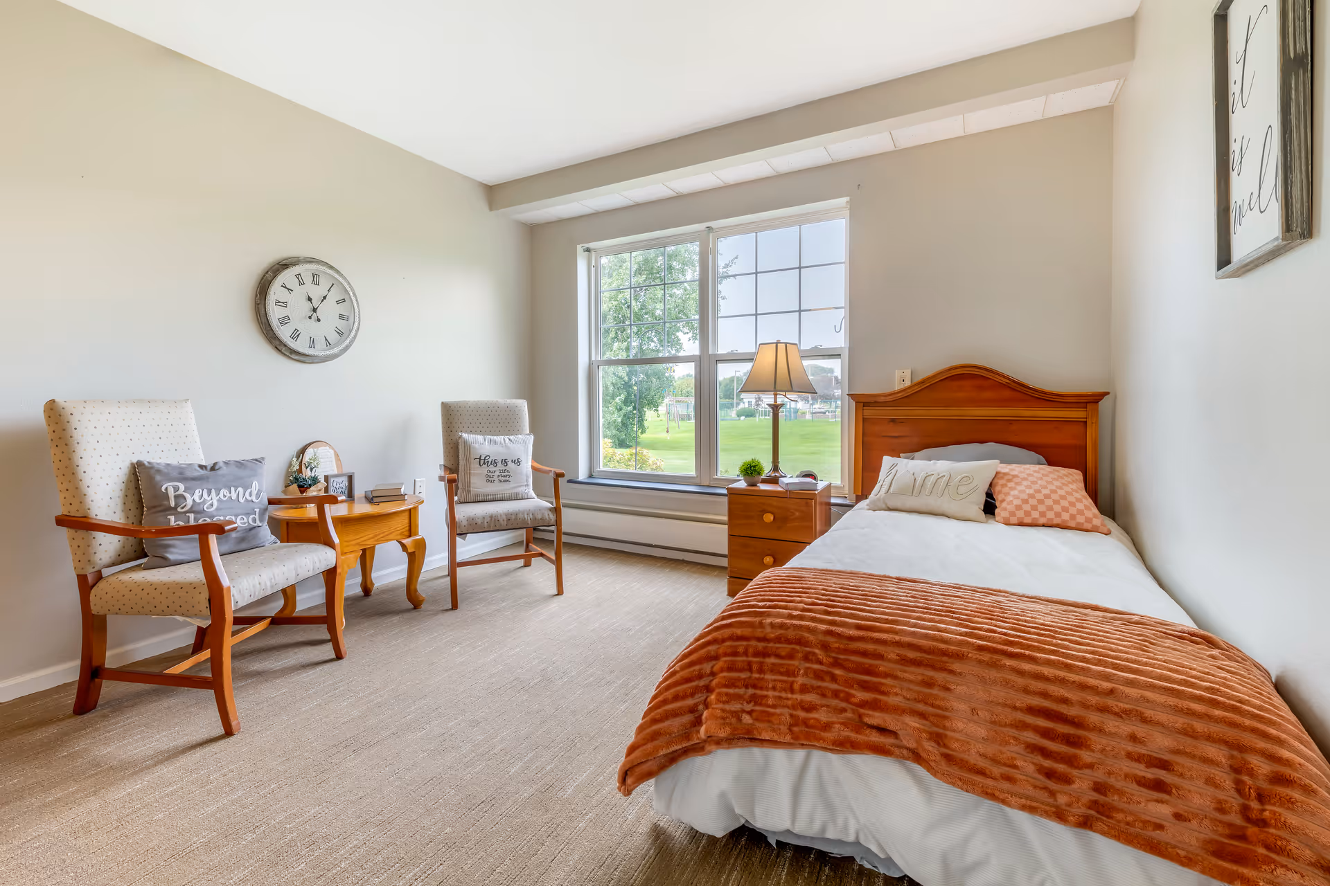 A bright and cozy bedroom with a single wooden bed covered with white bedding and an orange throw blanket. There are two cushioned armchairs with pillows, a small wooden side table between them, and a wooden nightstand with a lamp next to the bed. A large window lets in natural light and offers a view of green grass and trees outside. The walls are light-colored with a clock and framed artwork hanging on them.