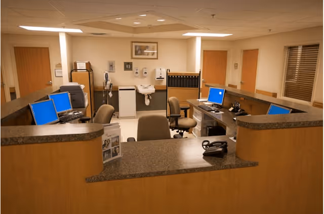 Reception or nurse station area with a U-shaped wooden counter, several office chairs, multiple computer monitors with blue screens, a telephone, and medical supplies mounted on the wall in the background.