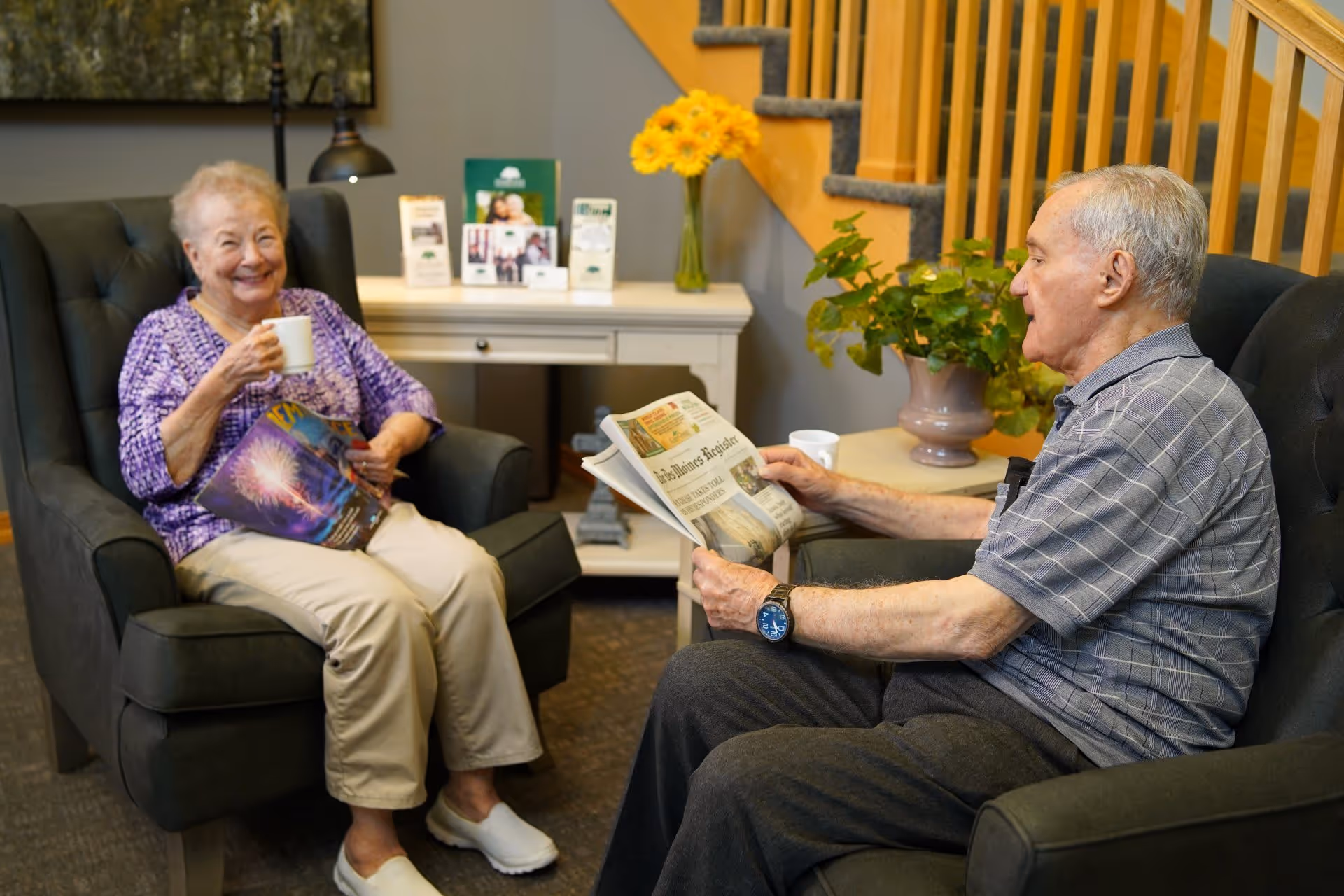 An elderly woman and man sitting in comfortable armchairs in a cozy living room. The woman is smiling and holding a white mug and a magazine, while the man is reading a newspaper. Behind them is a white console table with brochures, a lamp, and a vase with yellow flowers. A wooden staircase and a potted plant are visible in the background.