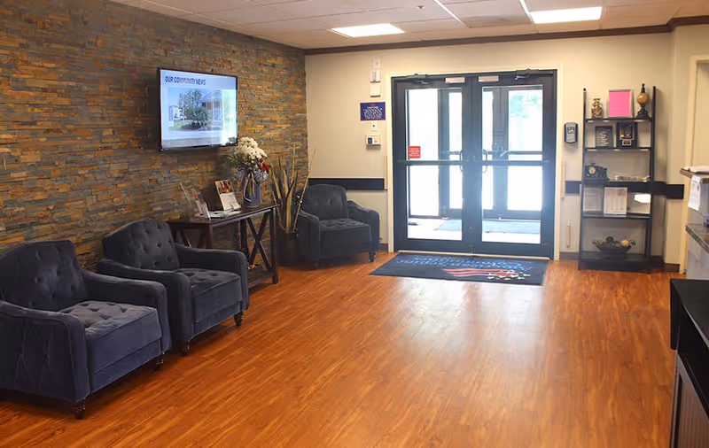 A waiting area with three dark blue upholstered armchairs arranged along a stone accent wall. A flat-screen TV is mounted on the wall above a small table with flowers and brochures. The floor is wood laminate, and there is a glass double door entrance with a welcome mat featuring an American flag design. A black shelving unit with decorative items and informational materials is positioned near the entrance.