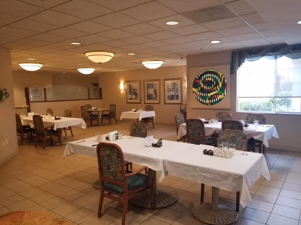 A dining room with several tables covered in white tablecloths, each set with glasses, napkins, and condiments. The chairs have patterned upholstery. The room has beige walls, tiled floors, ceiling lights, framed artwork on the walls, and a large window with a sheer curtain allowing natural light in.