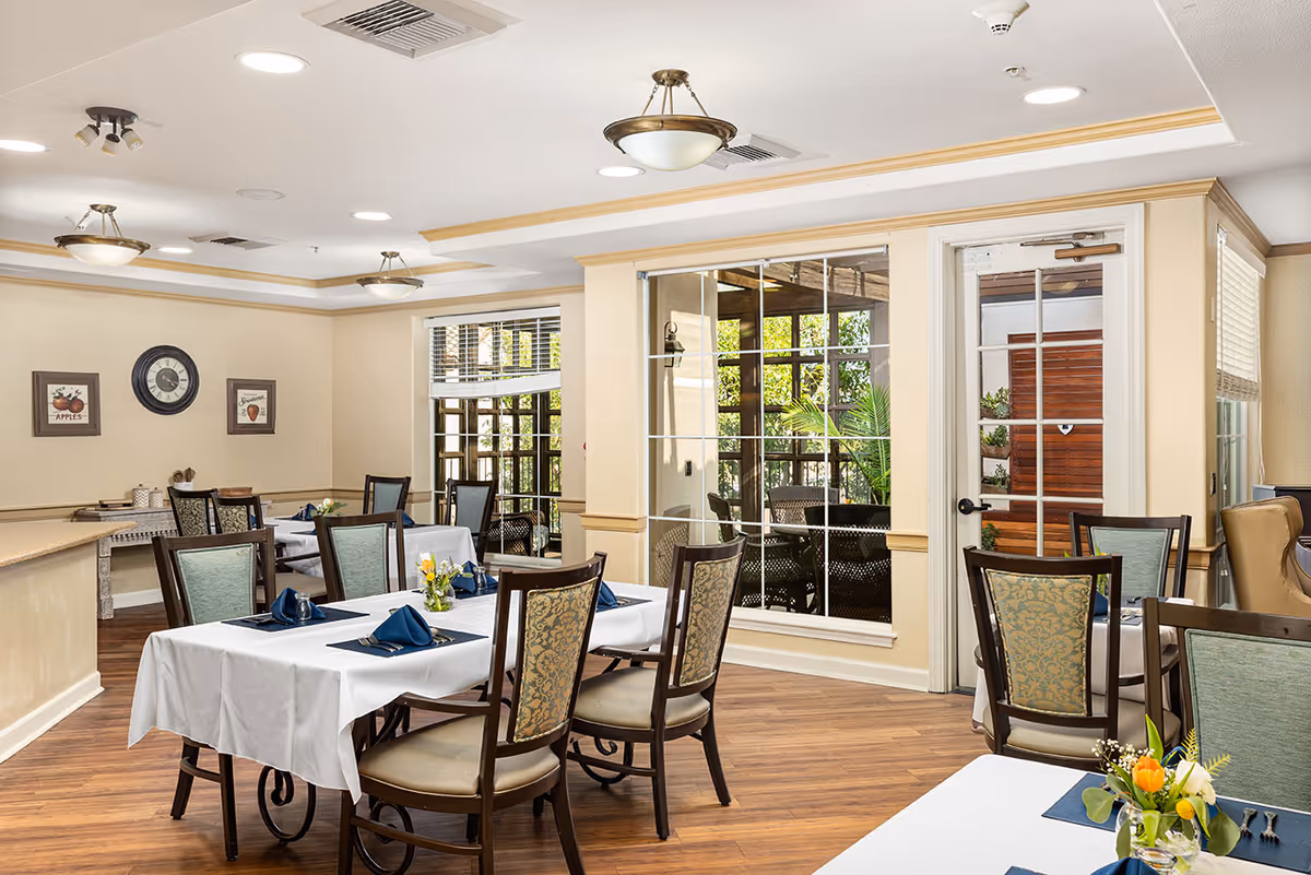 A bright and clean dining room with wooden floors and several tables covered with white tablecloths. Each table is set with blue napkins, silverware, and small floral centerpieces. The room has beige walls with framed artwork and a clock, ceiling lights, and large windows that look out onto a patio area with greenery.