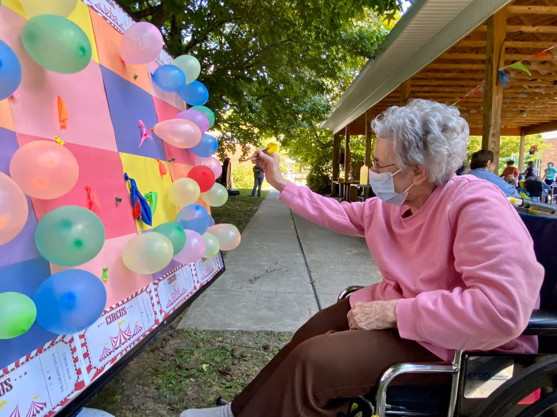 An elderly woman wearing a pink sweater and a face mask is sitting in a wheelchair outdoors, aiming a dart at a colorful balloon dart game board. The game board is decorated with balloons of various colors and has a circus theme. Other people and a covered patio area are visible in the background.