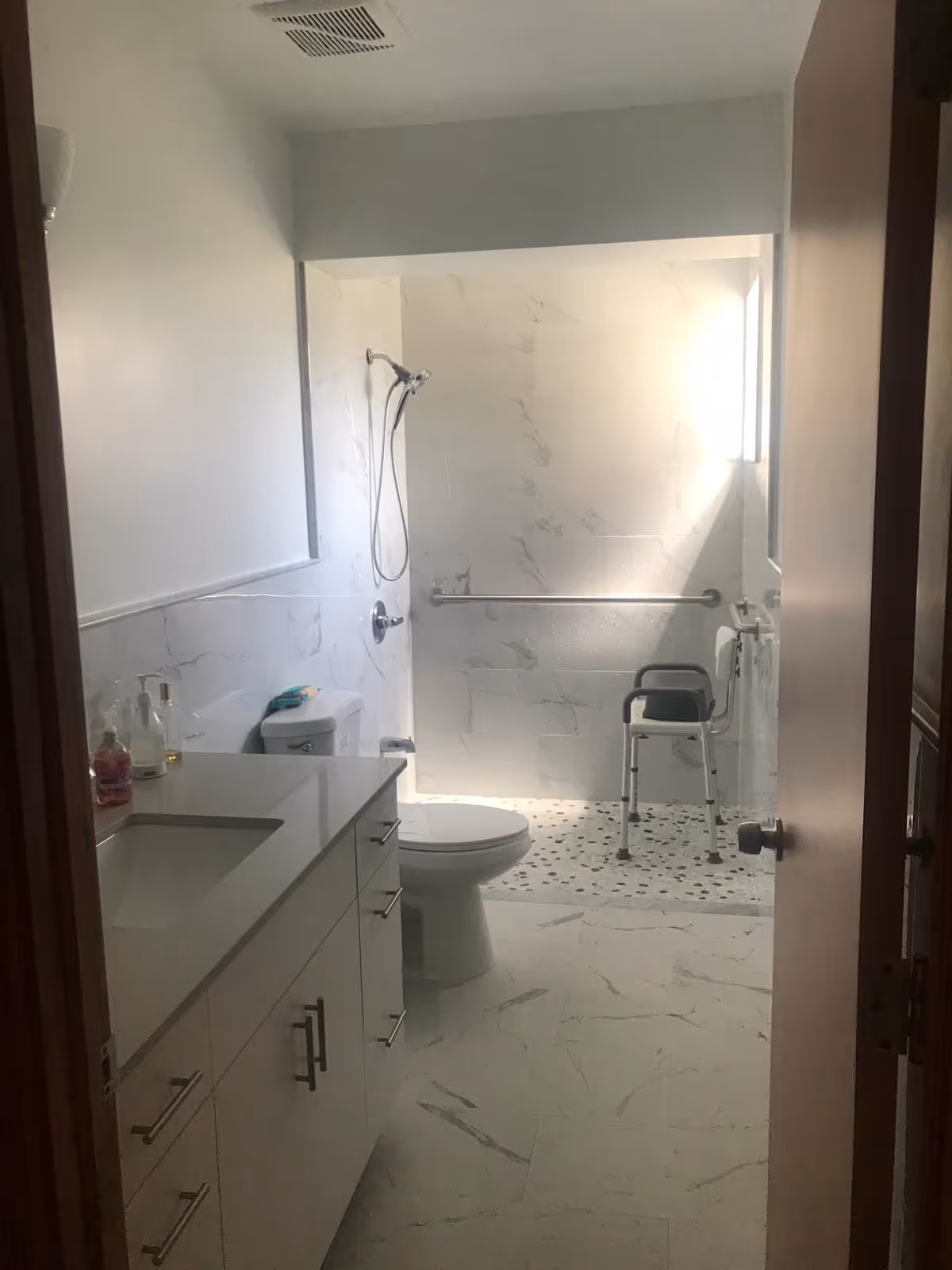 A bathroom with white marble-patterned tiles on the floor and walls. There is a white vanity with a sink and several drawers on the left side. A toilet is positioned near the center, and a walk-in shower area with a handheld showerhead and a shower chair is visible at the back. A grab bar is installed on the shower wall for support. Natural light is coming through a window in the shower area.