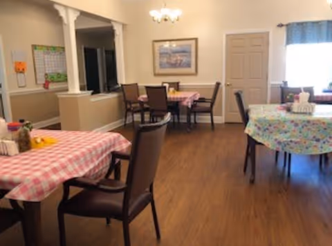 A dining room with several tables covered in colorful tablecloths, each surrounded by dark wooden chairs. The room has wooden flooring, beige walls, a chandelier, a framed picture on the wall, and a window with a blue valance letting in natural light.