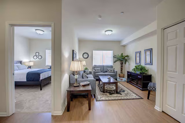 View of a cozy living room with two gray armchairs, a gray loveseat, a wooden coffee table on a patterned rug, a side table with a lamp, and framed artwork on the walls. To the left, there is an open doorway leading to a bedroom with a bed, nightstand, and lamp. The room has light-colored walls and wood flooring.