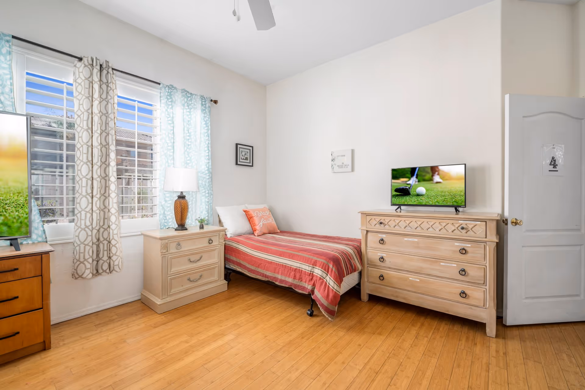 A bright and clean bedroom with a single bed covered in a red and beige striped bedspread, a wooden nightstand with a lamp and small plant, a wooden dresser with a TV on top displaying a golf scene, light blue and beige patterned curtains on the window, and a white door with a sign on it.