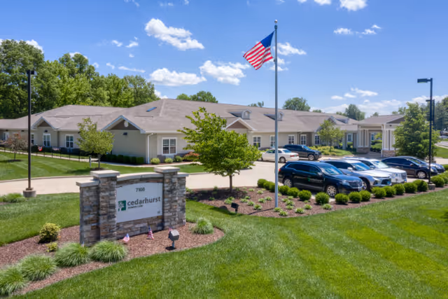 Exterior view of a single-story senior living facility building with a well-maintained lawn, several parked cars, an American flag on a flagpole, and a stone sign that reads '7108 cedarhurst memory care' under a clear blue sky with scattered clouds.