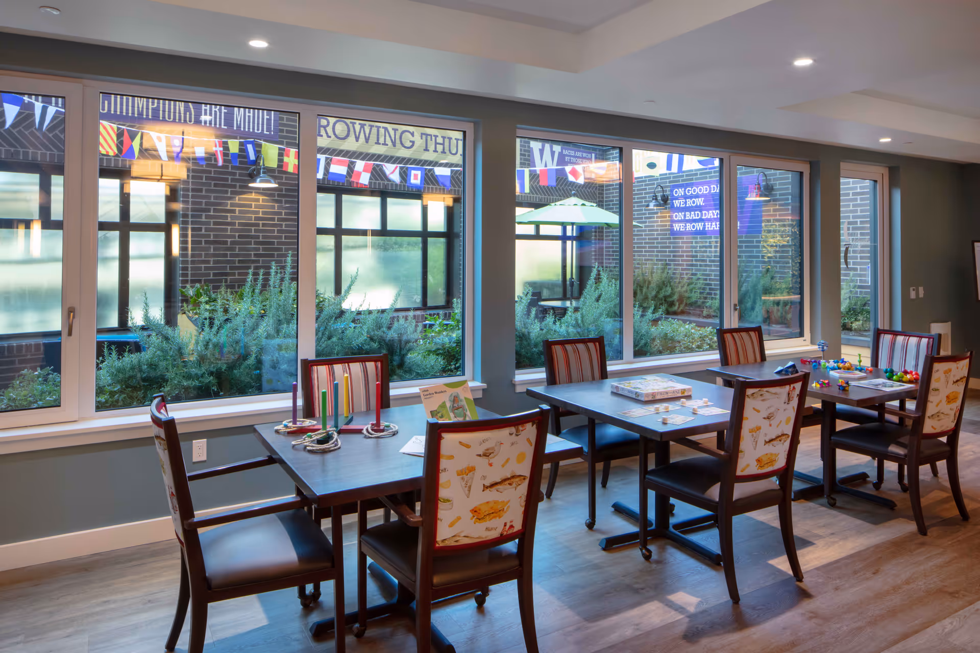 A bright room with large windows overlooking a garden area. Inside, there are several tables with chairs around them. The tables have various games and colorful objects on them. The chairs have patterned upholstery with images of food items. Outside the windows, there are decorative flags and signs on the brick walls.