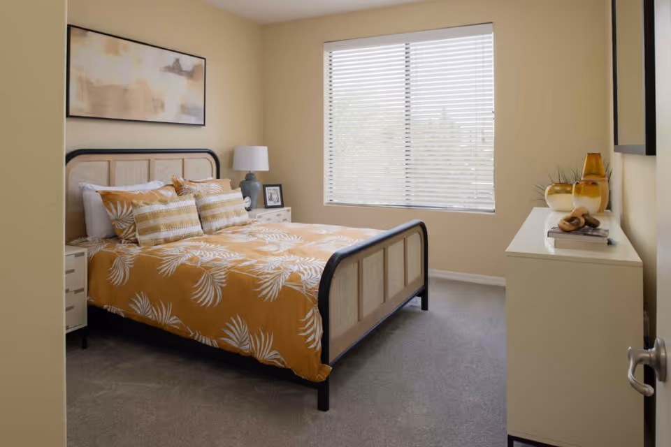 Bedroom with a bed dressed in orange leaf-patterned bedding, a bedside table with lamp, a dresser, and a window with blinds.