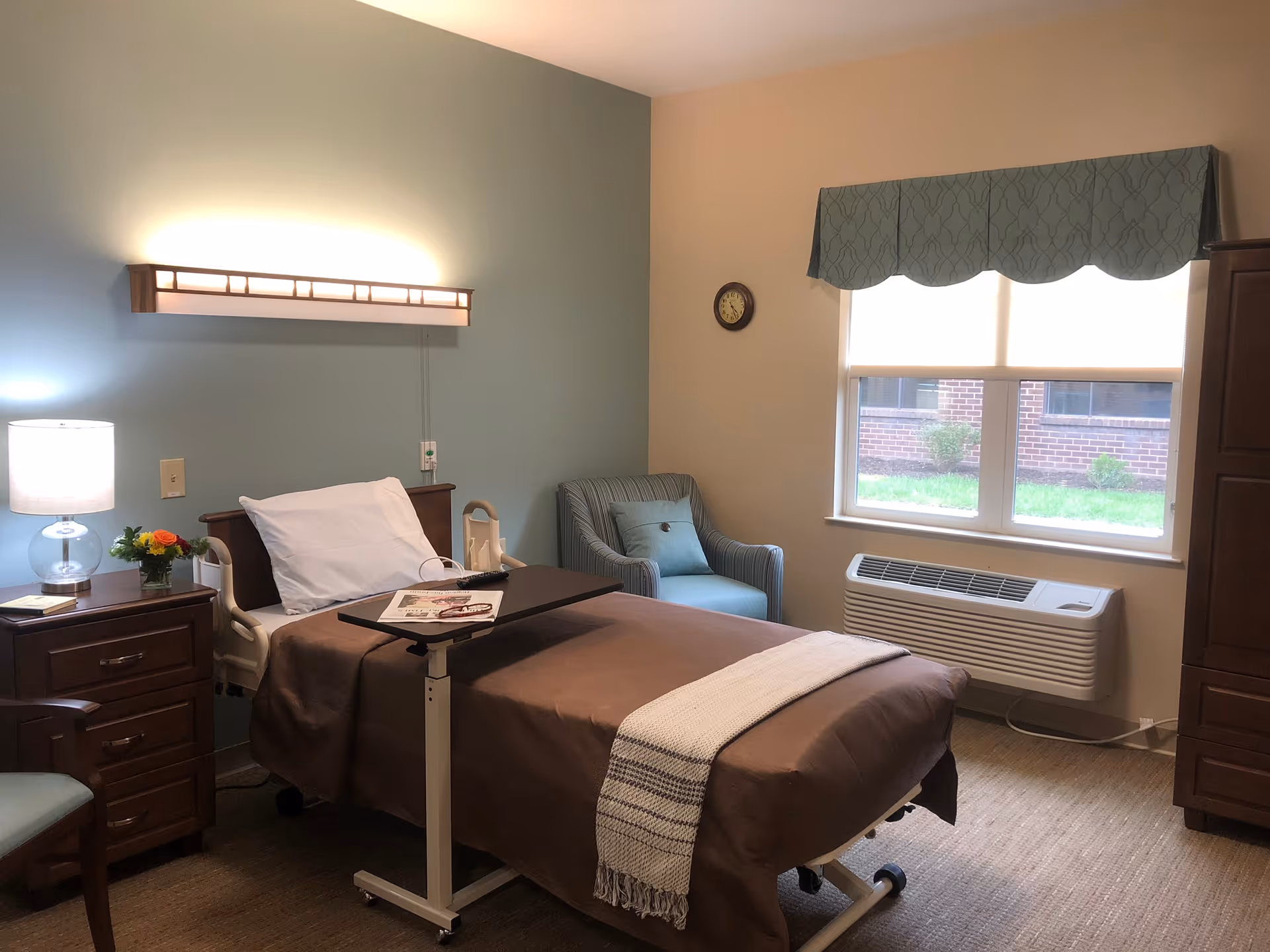 A neatly arranged bedroom in a senior living facility with a single hospital-style bed covered with a brown blanket and a white pillow. A small overbed table holds a magazine and a remote control. To the left of the bed is a wooden nightstand with a lamp, a small flower arrangement, and a book. A comfortable blue armchair is positioned near a window with a green valance, and a wall clock is mounted above the chair. The room has soft lighting and a calming color scheme with blue and beige walls.