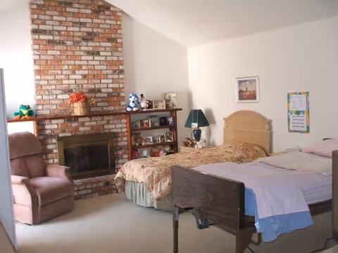 A bedroom with two beds, a recliner, brick fireplace, shelving with decorations, and a bedside lamp.