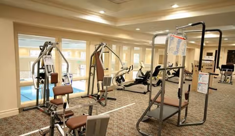 Indoor fitness room with various exercise machines including weight training equipment and stationary bikes. Large windows provide a view of an indoor swimming pool area.