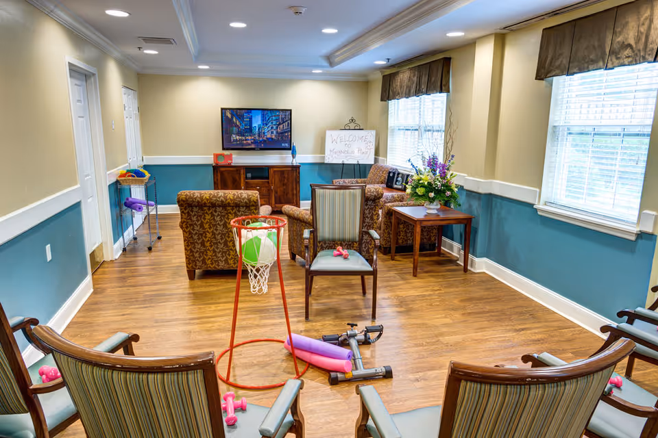 Cozy senior living common room with chairs arranged around a small exercise hoop, a television on a cabinet, and a welcome sign by the window.
