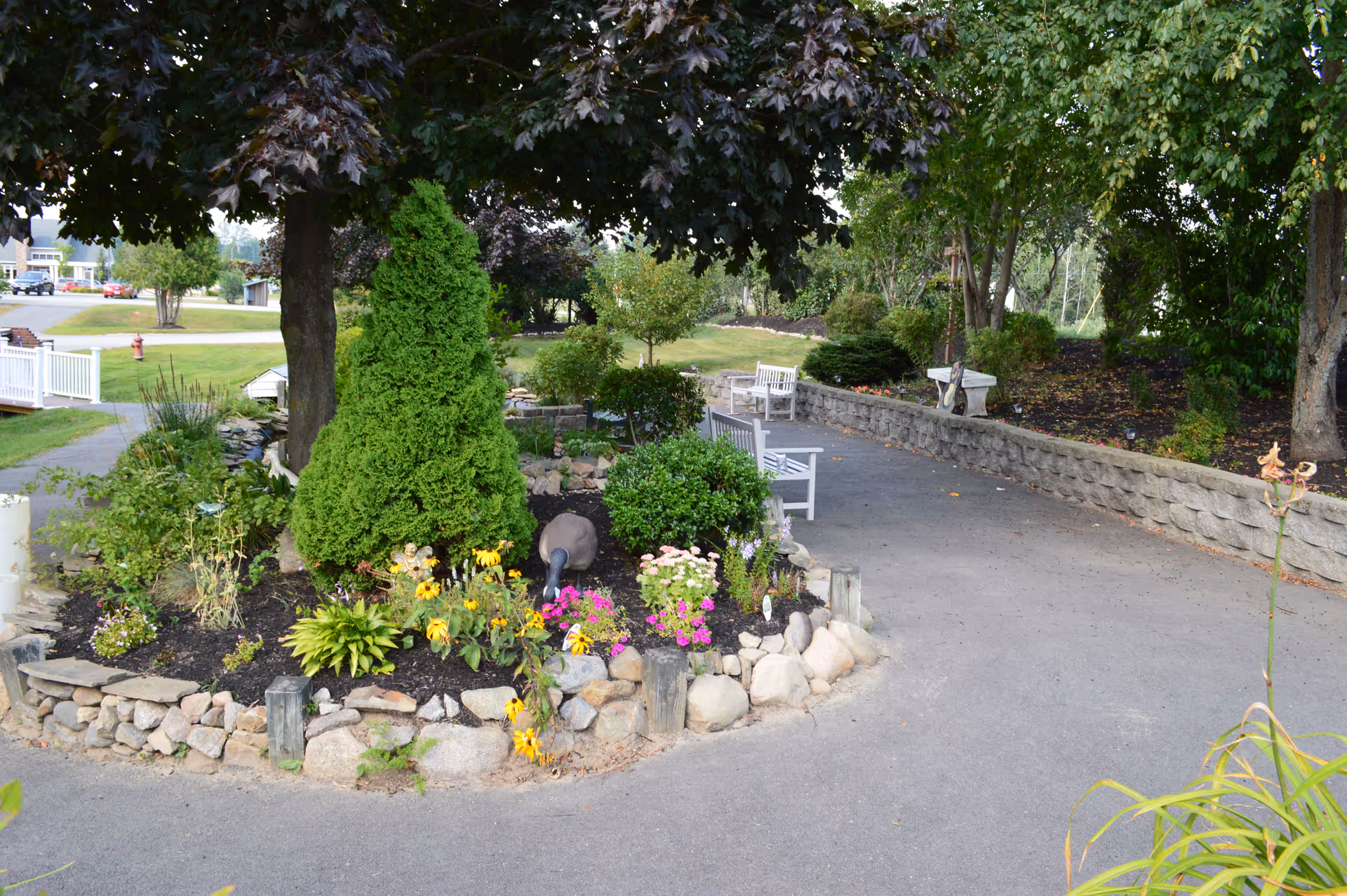 A landscaped outdoor garden area with a variety of green shrubs, flowering plants, and trees. There is a curved paved pathway with white benches along the side and a low stone wall bordering a garden bed. The setting appears peaceful and well-maintained.